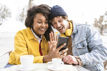 Happy woman with man waving on video call through smart phone at sidewalk cafe - OIPF01623