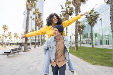 Happy woman with arms outstretched enjoying piggyback ride with man on footpath - OIPF01644