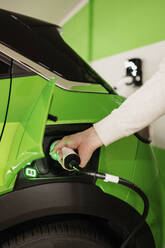 Hand of young man holding electric plug charging green car - GMCF00285