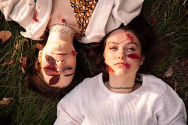 Zwei junge Frauen mit roten Blättern im Gesicht liegen im Herbst im Gras - CAVF96321