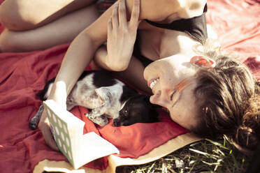 Beautiful woman smiles and reads to tiny puppy on an afternoon picnic - CAVF96398