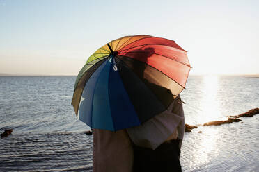 Lesbisches Paar küsst sich unter einem stolzen Regenschirm am Strand - CAVF96425