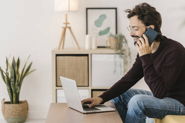 Glücklicher Mann mit Laptop, der zu Hause mit einem Mobiltelefon spricht - XLGF02908