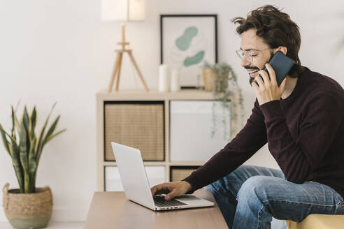 Happy man with laptop talking on mobile phone at home - XLGF02908