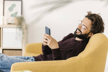 Happy man using smart phone sitting on sofa at home - XLGF02909