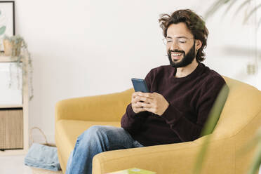 Happy man wearing eyeglasses using smart phone sitting on sofa at home - XLGF02911