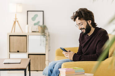 Smiling man wearing eyeglasses using smart phone sitting on sofa at home - XLGF02912