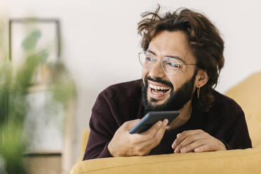 Glücklicher Mann mit Mobiltelefon auf dem Sofa liegend zu Hause - XLGF02914