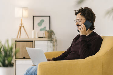 Smiling man with laptop talking on mobile phone at home - XLGF02916