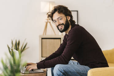 Lächelnder bärtiger Hipster-Mann mit Laptop, der zu Hause auf dem Sofa sitzt - XLGF02918