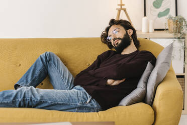 Thoughtful bearded man with arms crossed relaxing on sofa at home - XLGF02921