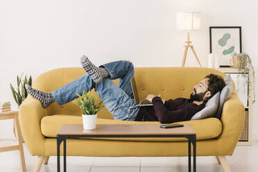 Mann mit Laptop auf dem Sofa im Wohnzimmer zu Hause liegend - XLGF02925