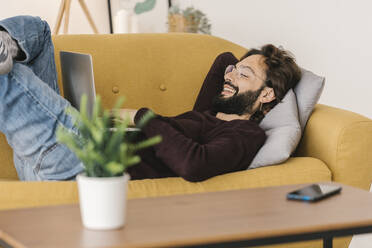 Lächelnder Mann mit Laptop auf dem Sofa im Wohnzimmer zu Hause liegend - XLGF02926