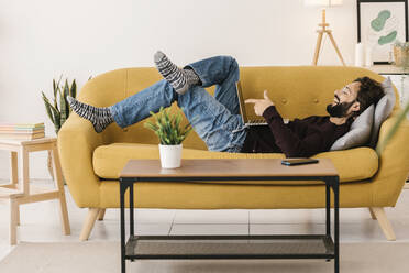 Happy man using laptop for video call lying on sofa at home - XLGF02927