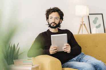 Thoughtful bearded man with tablet PC sitting on sofa in living room at home - XLGF02929