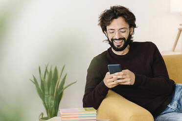 Glücklicher Mann mit Bart, der ein Mobiltelefon benutzt und zu Hause auf dem Sofa sitzt - XLGF02931