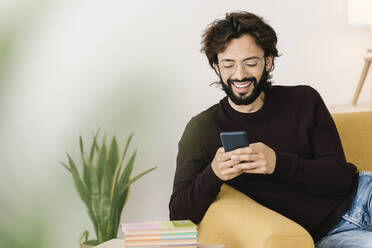 Happy man with beard using mobile phone sitting on sofa at home - XLGF02931