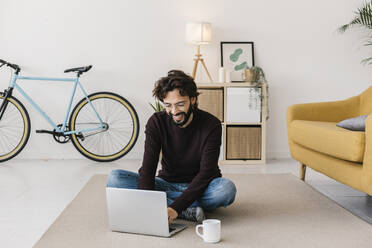 Glücklicher Mann mit Laptop auf Teppich sitzend zu Hause - XLGF02935