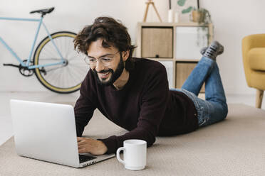 Lächelnder Mann, der einen Laptop benutzt, der neben einer Kaffeetasse auf dem Teppich zu Hause liegt - XLGF02937