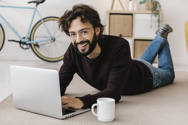 Lächelnder Mann mit Laptop und Kaffeetasse auf dem Teppich im Wohnzimmer liegend zu Hause - XLGF02938