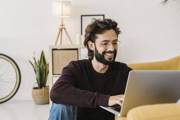 Smiling bearded man using laptop sitting by sofa at home - XLGF02943