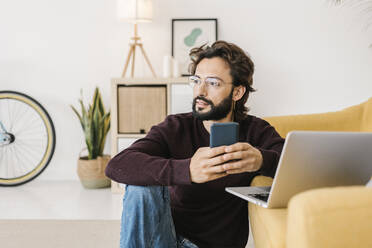 Nachdenklicher bärtiger Mann mit Mobiltelefon und Laptop, der zu Hause auf dem Sofa sitzt - XLGF02947