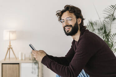 Glücklicher Mann mit Mobiltelefon im Wohnzimmer zu Hause - XLGF02952