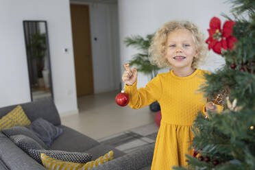 Lächelndes blondes Mädchen mit Christbaumkugel im Wohnzimmer stehend - SVKF00131