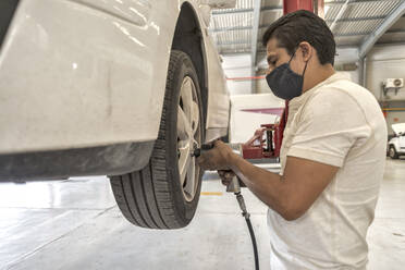 Latin male mechanic removing a white car's wheel with an impact wrench - CAVF96517