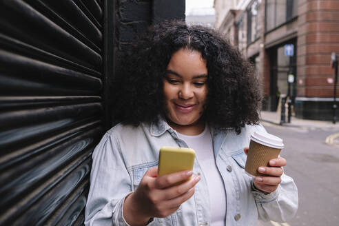 Lächelnde junge Frau, die einen Einweg-Kaffeebecher hält und ihr Smartphone mit einem Auslöser benutzt - ASGF02268
