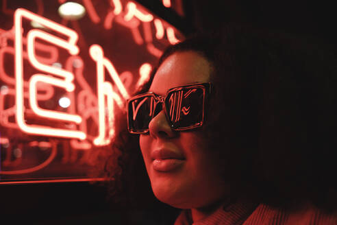 Junge Frau mit Sonnenbrille an einer beleuchteten Wand bei Nacht - ASGF02288