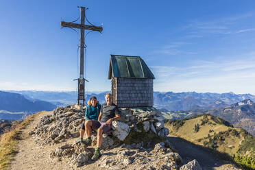 Deutschland, Bayern, Zwei Wanderer beim Ausruhen vor dem Gipfelkreuz auf dem Geigelstein - FOF13132