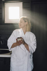 Woman in bathrobe holding cup enjoying sunlight at home - VPIF05919