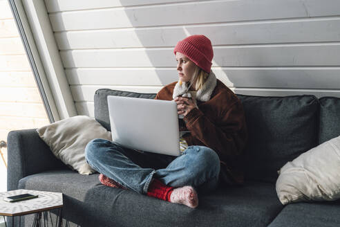 Frau in warmer Kleidung sitzt im Schneidersitz auf dem Sofa mit Laptop zu Hause - VPIF05945
