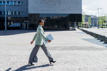 Selbstbewusste Geschäftsfrau mit Laptop, die an einem Bürogebäude vorbeigeht - MEUF05224