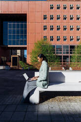 Junge Geschäftsfrau tippt auf einem Laptop vor einem Bürogebäude - MEUF05243