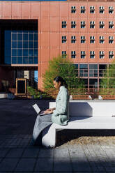 Young businesswoman typing on laptop by office building - MEUF05243