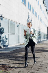 Junge Geschäftsfrau mit Smartphone, die auf einem Fußweg an einem Bürogebäude vorbeigeht - MEUF05258