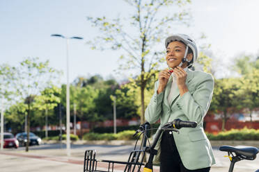 Businesswoman with electric bicycle adjusting cycling helmet on street - MEUF05265