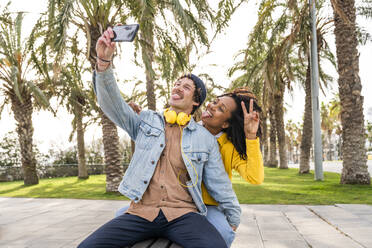 Happy couple sticking out tongues taking selfie through mobile phone at park - OIPF01651