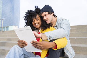 Happy woman sharing tablet PC with boyfriend sitting on steps - OIPF01667