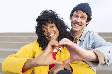 Happy couple gesturing heart shape sitting together on steps - OIPF01669