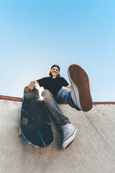 Happy man with skateboard sitting at skateboard park - OMIF00756