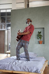 Älterer Mann mit Kopfhörern, der zu Hause auf dem Bett Gitarre spielt - FMKF07460