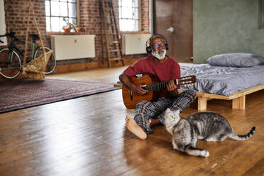 Mature man playing guitar by cats in bedroom at home - FMKF07465