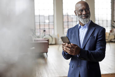 Smiling businessman with mobile phone at home - FMKF07494