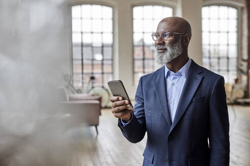 Thoughtful businessman with mobile phone at home - FMKF07495