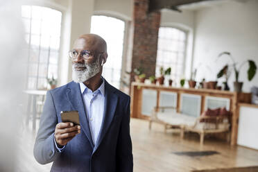 Contemplative businessman with smart phone in living room - FMKF07497