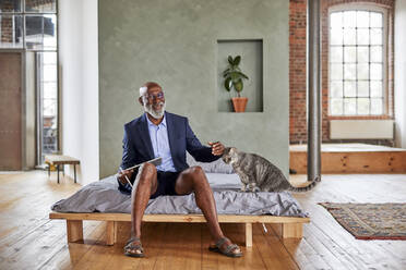 Smiling businessman with tablet computer stroking cat on bed in bedroom - FMKF07557