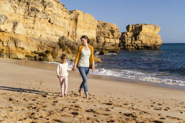 Mother and daughter walking in front of rock formation on shore at beach - DIGF17850